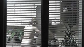 Peeping. Voyeur. Neighbors pervert voyeur inside evening on street looks out window as nude beauty neighbors inside kitchen prepares dinner for her hubby. Nude into outdoor. Nude at home. Family. Public