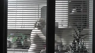 Peeping. Voyeur. Neighbors pervert voyeur inside evening on street looks out window as nude beauty neighbors inside kitchen prepares dinner for her hubby. Nude into outdoor. Nude at home. Family. Public