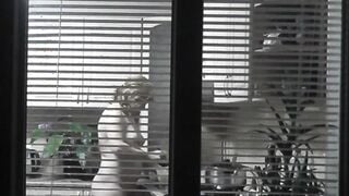 Peeping. Voyeur. Neighbors pervert voyeur inside evening on street looks out window as nude beauty neighbors inside kitchen prepares dinner for her hubby. Nude into outdoor. Nude at home. Family. Public