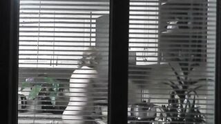 Peeping. Voyeur. Neighbors pervert voyeur inside evening on street looks out window as nude beauty neighbors inside kitchen prepares dinner for her hubby. Nude into outdoor. Nude at home. Family. Public