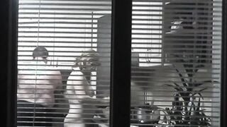 Peeping. Voyeur. Neighbors pervert voyeur inside evening on street looks out window as nude beauty neighbors inside kitchen prepares dinner for her hubby. Nude into outdoor. Nude at home. Family. Public