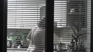 Peeping. Voyeur. Neighbors pervert voyeur inside evening on street looks out window as nude beauty neighbors inside kitchen prepares dinner for her hubby. Nude into outdoor. Nude at home. Family. Public