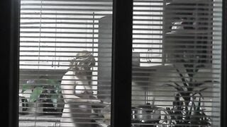 Peeping. Voyeur. Neighbors pervert voyeur inside evening on street looks out window as nude beauty neighbors inside kitchen prepares dinner for her hubby. Nude into outdoor. Nude at home. Family. Public